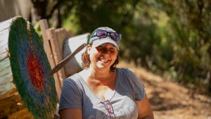 A smiling woman in a gray shirt and cap stands in front of an archery target with an arrow sticking out, appearing as though it went through her head due to the angle. She is outdoors with trees in the background.