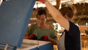 Two women work together outdoors, one holding open the lid of a large blue chest freezer while the other, wearing gloves and safety glasses, handles an object inside. Sunlight filters through a rustic wooden shelter.