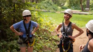 Two women wearing helmets and harnesses smile and talk outdoors on a forest path, enjoying a sunny day surrounded by greenery. Another person in a helmet is partially visible in the foreground.