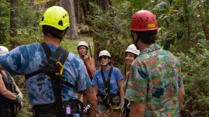 A group of people wearing helmets and harnesses stand together in a forest, preparing for a ziplining or ropes course activity. Tall trees and green foliage surround them.