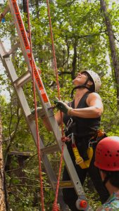 A person wearing a helmet and harness climbs a ladder secured with ropes in a wooded area, while another helmeted person stands nearby. Sunlight filters through the trees above.