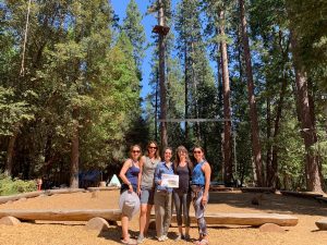 Five women stand together outdoors in a wooded area with tall trees, sunlight filtering through. They are smiling at the camera, and one is holding a white sign. Wood chips cover the ground. A ropes course is visible in the background.