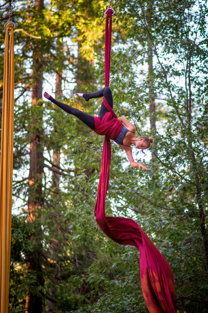 A female aerialist performs on red silks, suspended high in the air among tall trees. She is mid-pose, with one leg wrapped in the fabric and her body arched gracefully against a forest backdrop.