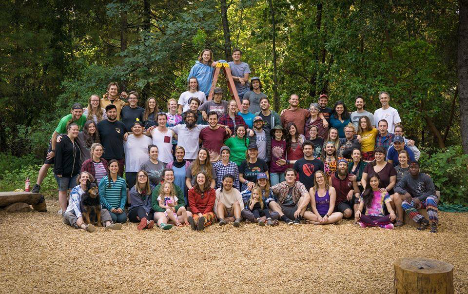 A large group of people of various ages and backgrounds pose together outdoors in a wooded area, smiling and sitting or standing in rows on wood chips, with trees and greenery in the background.