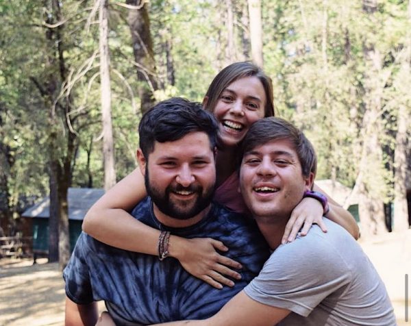 Three young adults smile and laugh together outdoors in a wooded area, reminiscing about the best summer camp food. One woman hugs two men from behind as they enjoy the natural setting, with sunlight filtering through tall trees in the background.