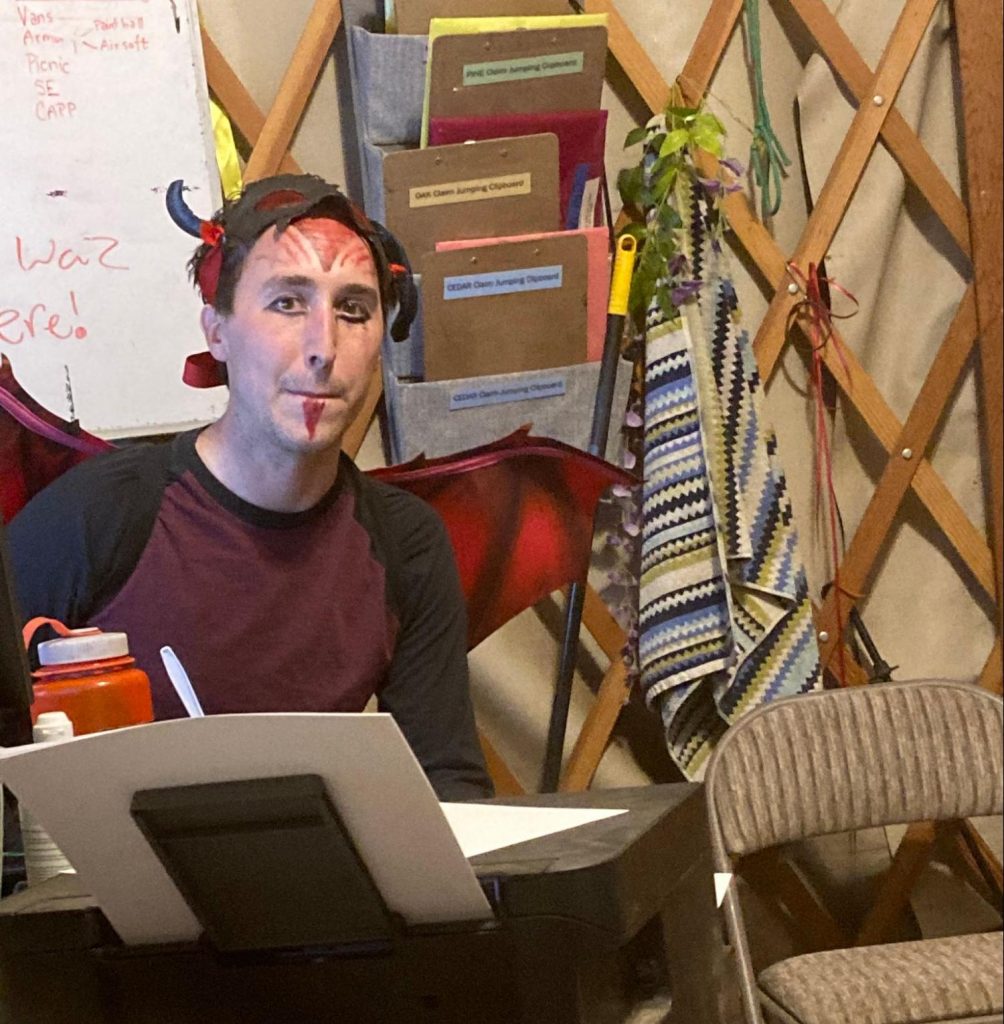 A person with red face paint, horn headband, and stick-on goatee sits at a keyboard in a cluttered room with folders, a striped blanket, and notes on the wall behind them.