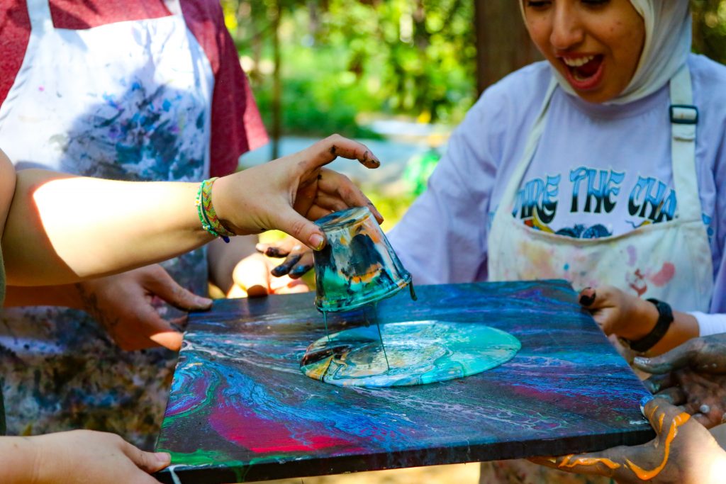 Three people enjoy family camp activities outdoors, using the paint pouring technique on a canvas. One person lifts a paint-covered cup, another holds the canvas, while a smiling woman in a paint-splattered apron and hijab looks on.