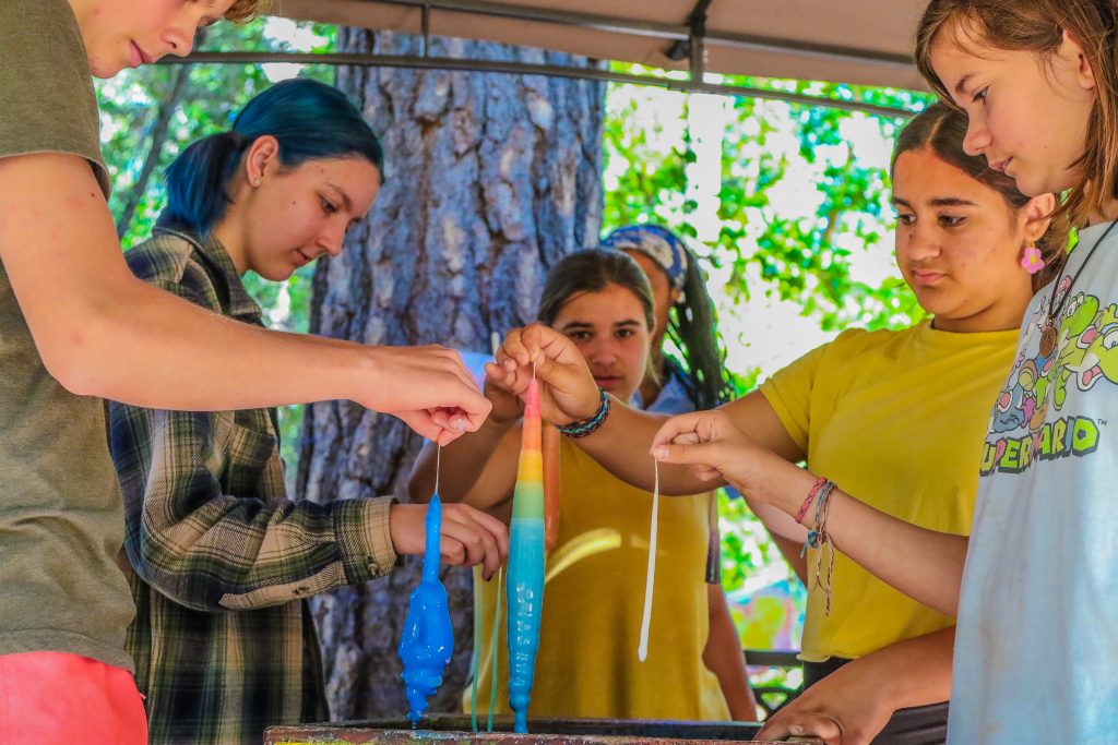 A group of young people stand around a table, dipping long white strings into colored wax to make candles outdoors, surrounded by trees and sunlight—a classic scene from family camp activities.