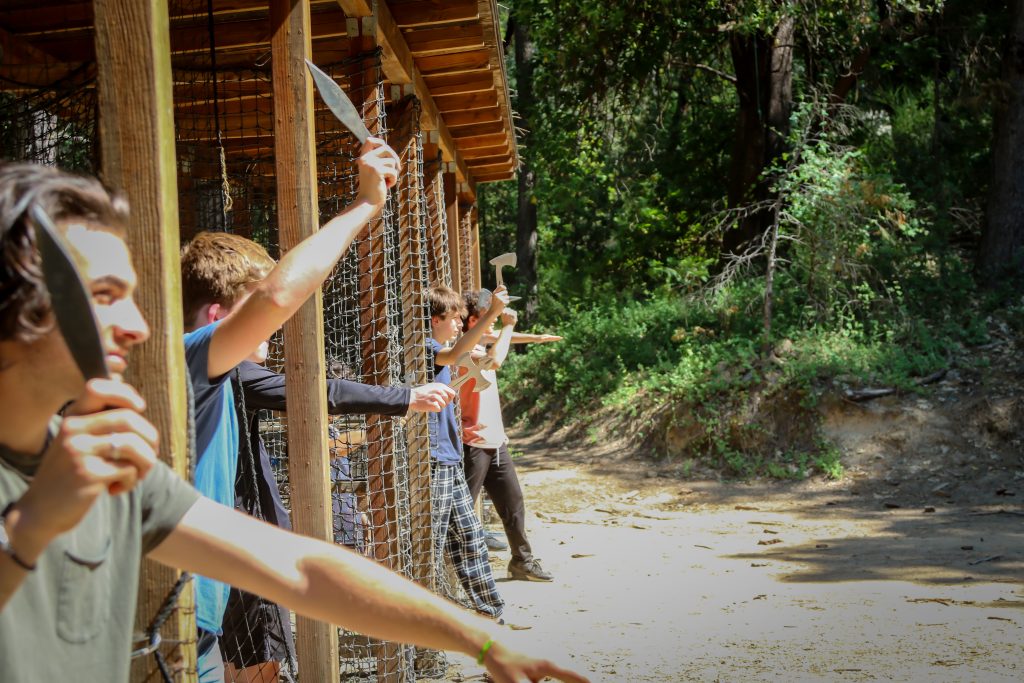 Four people stand in a row at an outdoor range, each holding a knife or hatchet ready to throw at targets ahead. Trees and sunlight fill the background.