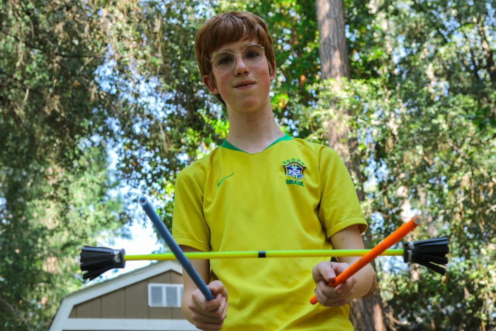 A boy wearing glasses and a yellow Brazil soccer jersey stands outdoors, holding three toy arrows with rubber tips—perfect for family camp activities. Trees and a shed are visible in the background.