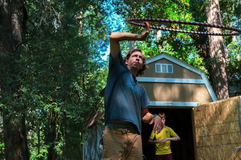 A man stands outside near a shed, spinning a hula hoop above his head with one hand. Sunlight filters through the trees as family camp activities unfold, with another person in a yellow shirt visible in the background.