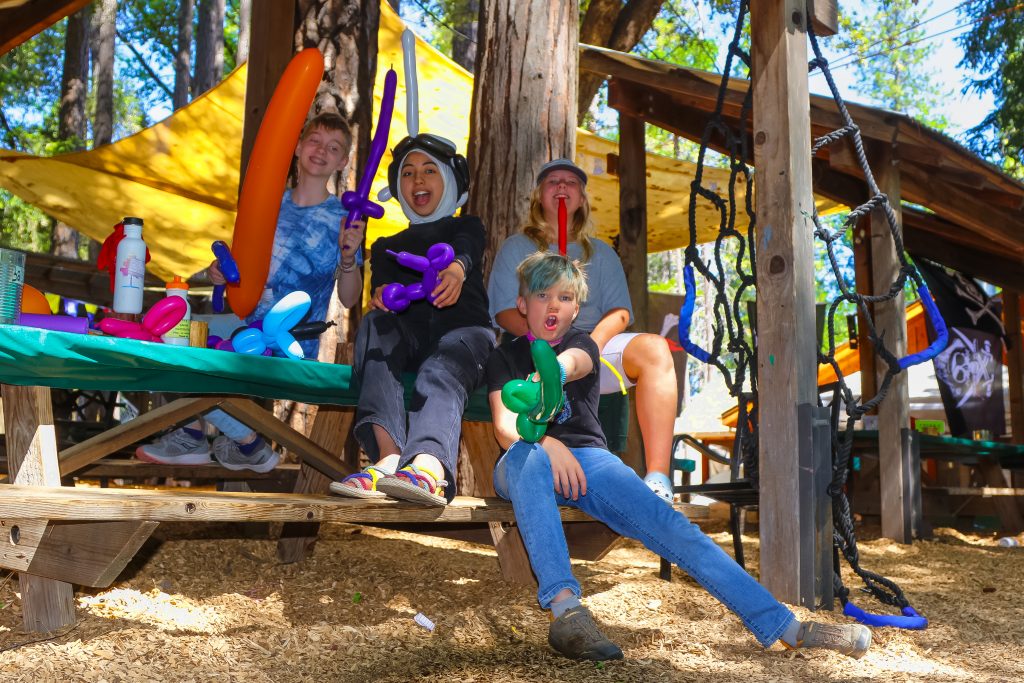 Four people sitting on wooden benches outside, playfully posing with colorful balloon swords and animals. Surrounded by trees and playful decorations, they appear to be enjoying family camp activities in a lively, fun-filled setting.