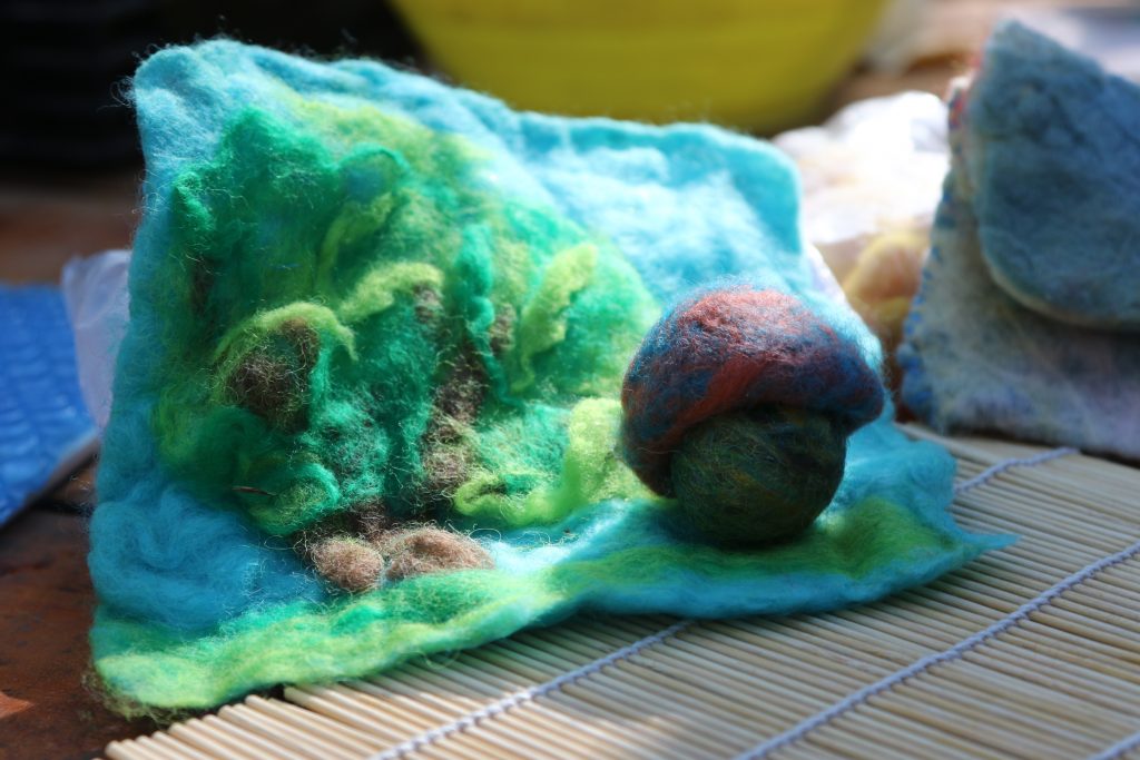 A piece of blue and green felted wool with textured patterns lies on a bamboo mat, next to a round, multicolored felted wool ball—perfect inspiration for family camp activities. A yellow bowl and other felt pieces are blurred in the background.