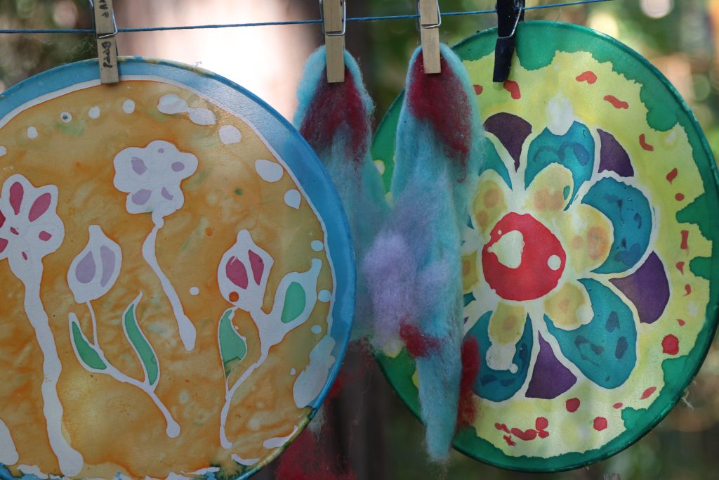 Two circular, colorful batik art pieces with floral designs hang on a clothesline with clothespins—perfect for family camp activities. Fluffy, dyed wool in red, blue, and white hangs between them as sunlight filters through leafy branches.