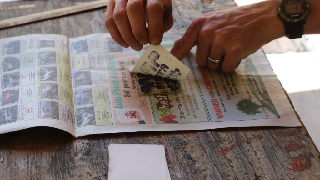 A person presses a small card with black ink onto a newspaper on a worn wooden table, possibly making a print or stamp as part of creative family camp activities. Only their hands and part of their wristwatch are visible.