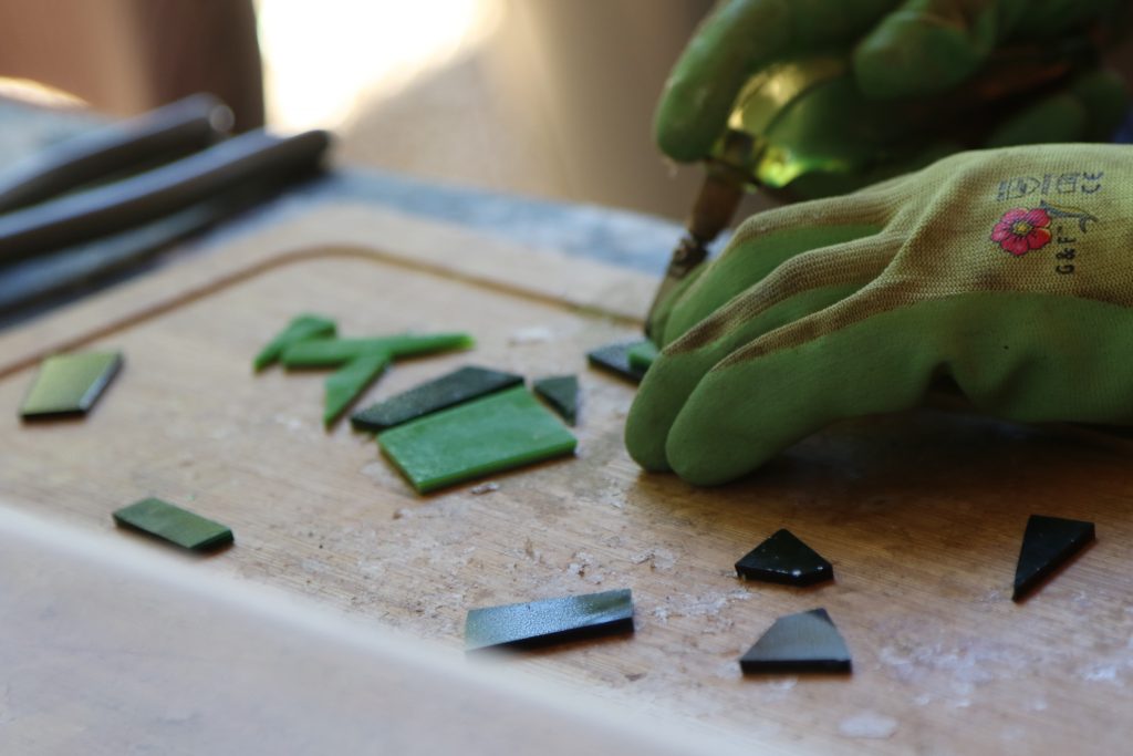 A person wearing green gloves uses a tool to work with small pieces of green and black stained glass on a wooden surface, demonstrating creative skills often enjoyed during family camp activities, with other tools visible in the background.