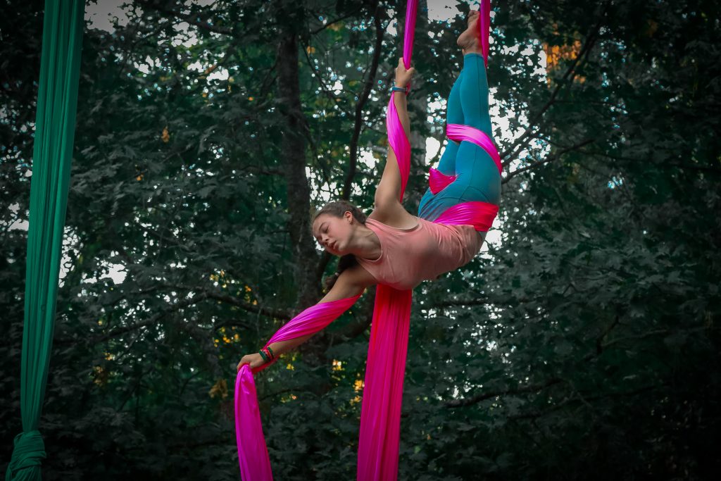 A woman performs an aerial silk routine outdoors, suspended by pink fabric, with green trees in the background. She is wearing a pink top and turquoise leggings, balancing gracefully in mid-air.