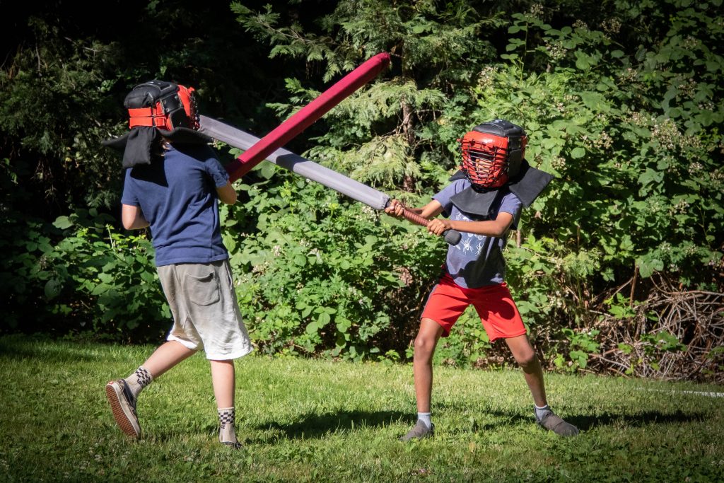 Two children wearing helmets and protective gear playfully duel with foam swords outside on a grassy area, surrounded by green bushes and trees—perfect for family camp activities.
