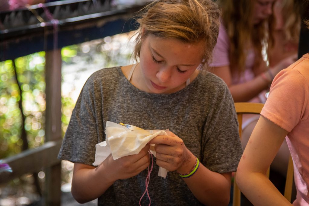 A young person with light brown hair and a gray shirt is focused on embroidering fabric with pink thread, sitting at a table outdoors during family camp activities. Other children are blurred in the background, also engaged in crafts.