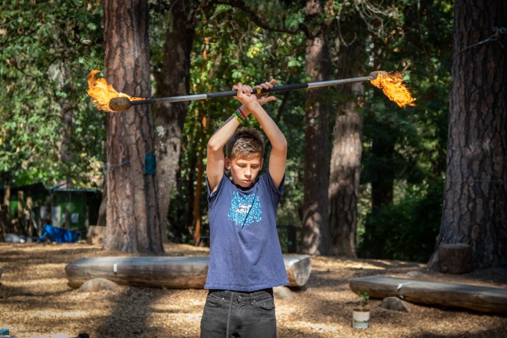 A young boy outdoors holds a flaming staff above his head with both arms crossed, concentrating as he performs in a wooded area with large trees and wooden benches in the background.