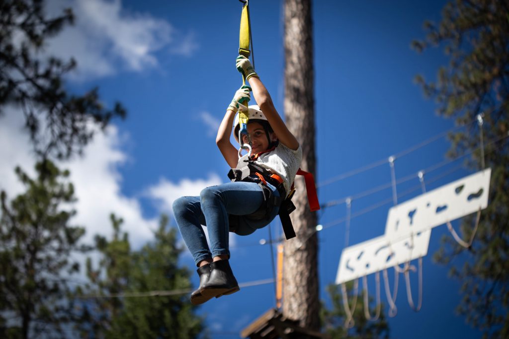 A child wearing a helmet and harness glides down a zip line in an outdoor adventure park, with a forest background and blue sky visible.