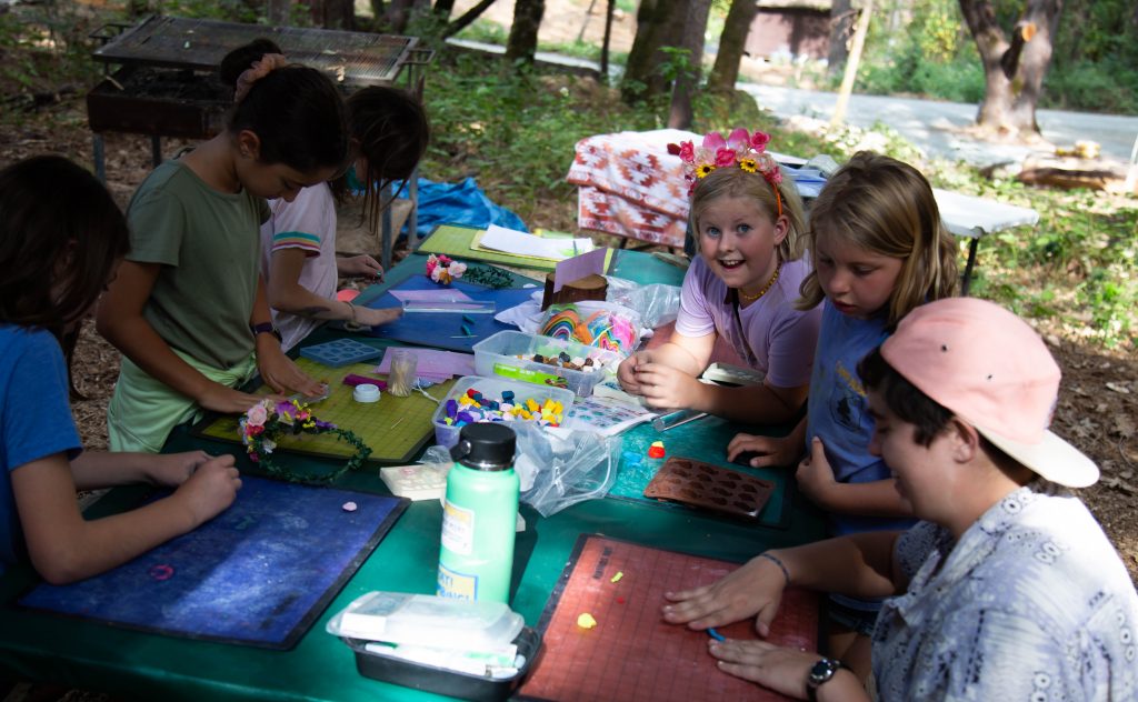 Children sit around a picnic table outdoors, engaging in family camp activities and crafts with colorful materials. One girl with a flower crown smiles at the camera while others focus on their projects. Trees and sunlight surround the scene.