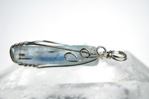 A blue crystal wrapped in intricate silver wire, forming a pendant, is displayed on a clear quartz surface with a bright, blurred background.