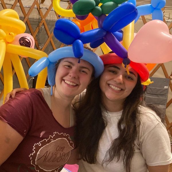 Two smiling young women stand close together wearing colorful balloon hats, one with blue and purple balloons and one with red, yellow, and a pink heart-shaped balloon, inside a room with a wooden lattice wall.