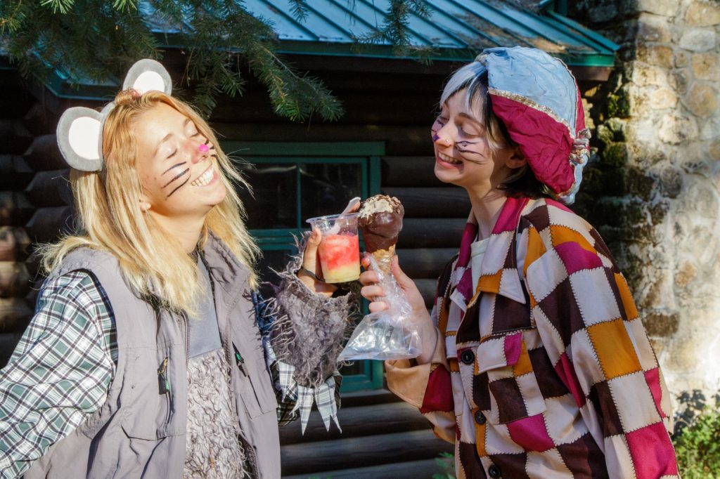 Two people in colorful costumes with face paint resembling mice smile and hold up frozen treats outside a log cabin, enjoying a sunny day.