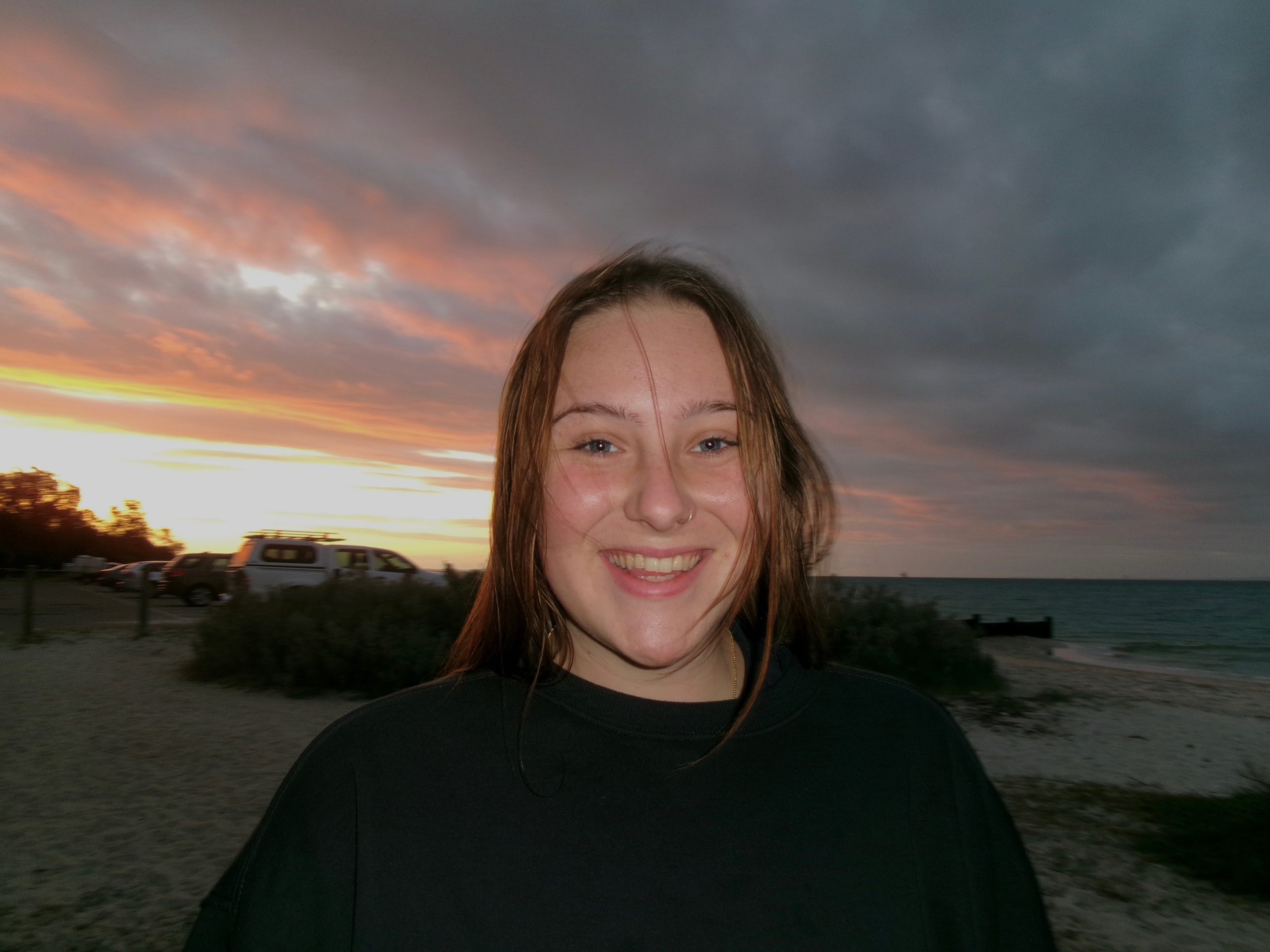A young woman with long brown hair smiles at the camera on a beach at sunset, with sand, greenery, cars, and the ocean in the background under a cloudy sky with orange and pink hues.