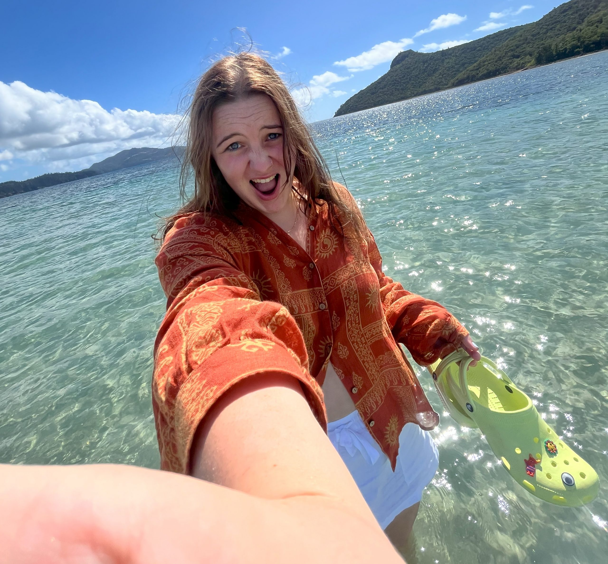 A young woman standing in clear, shallow water at the beach, wearing a red patterned shirt and white shorts, smiling and holding a yellow sandal. Green hills and a blue sky are visible in the background.