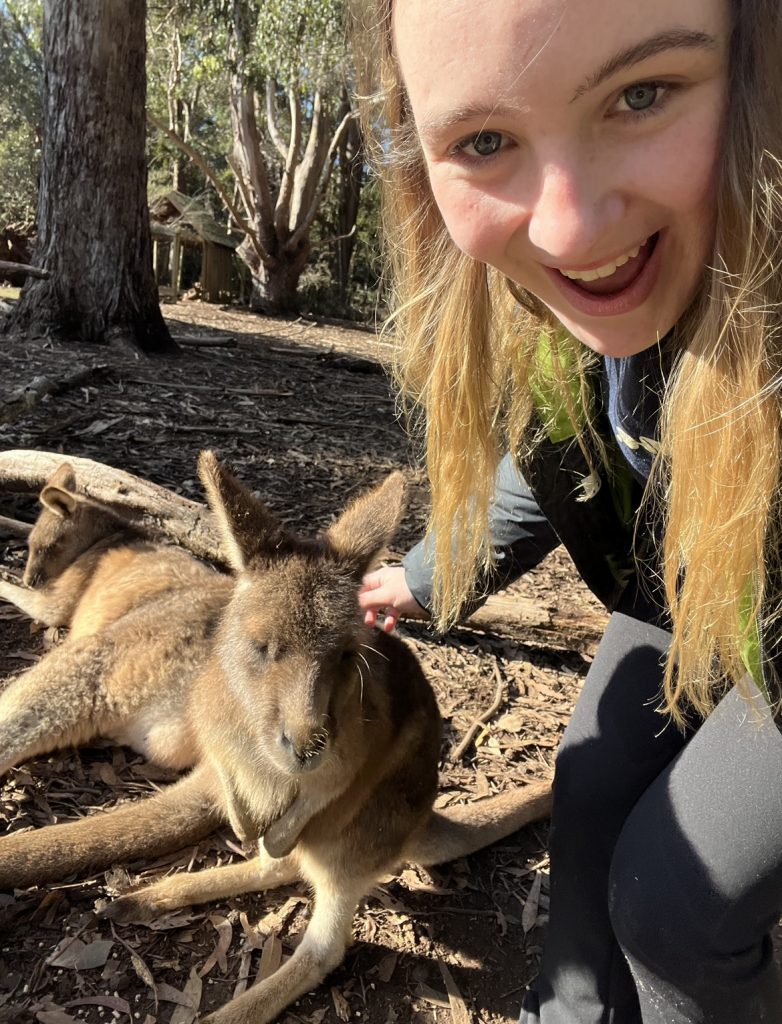 A smiling person takes a selfie while petting a relaxed kangaroo lying on the ground in a sunlit wooded area, with another kangaroo resting nearby.