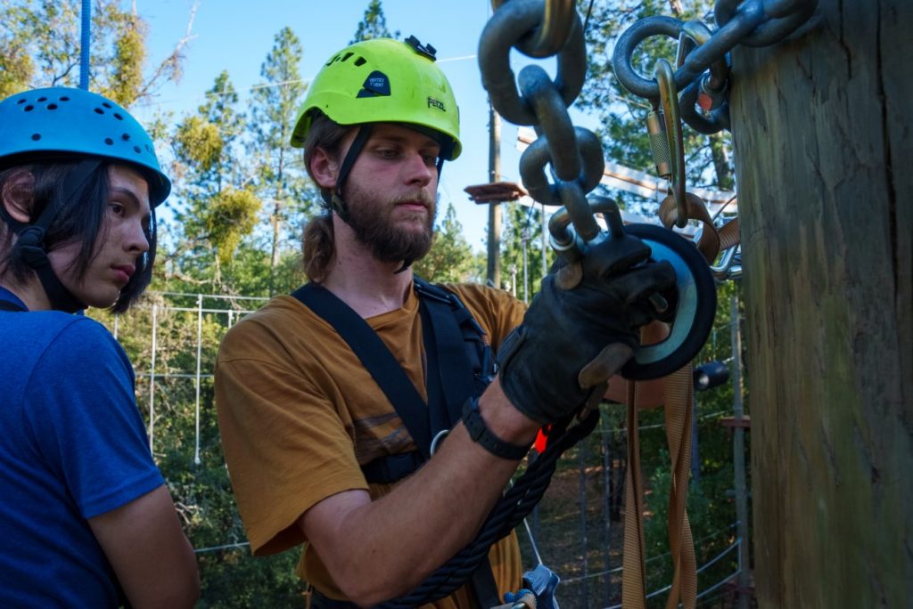 Two people wearing helmets and harnesses stand by a wooden pole outdoors. One is adjusting climbing equipment and carabiners while the other watches. Trees and ropes can be seen in the background.