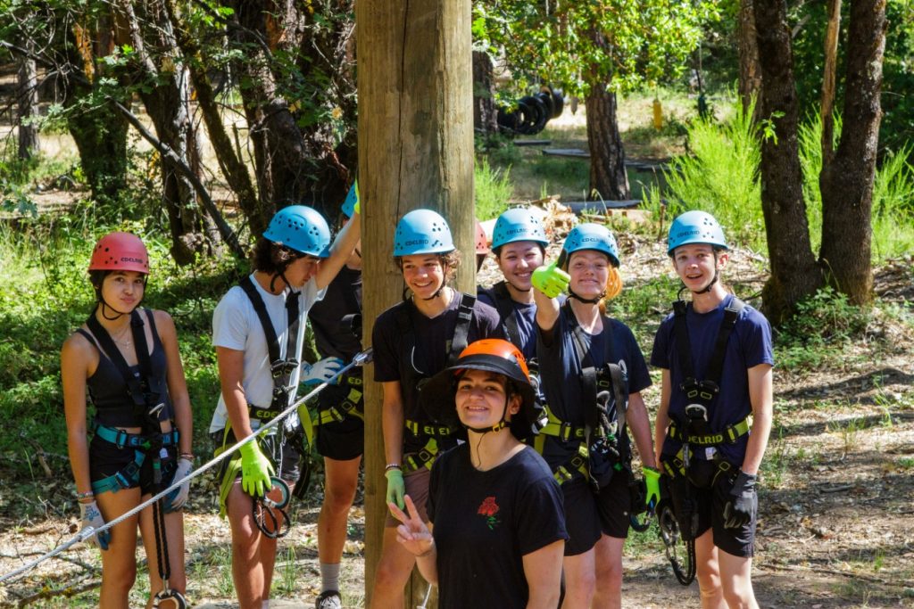 A group of seven people wearing helmets, harnesses, and gloves stand together outdoors in a wooded area, smiling at the camera. One person in front flashes a peace sign. Ropes and climbing gear are visible.