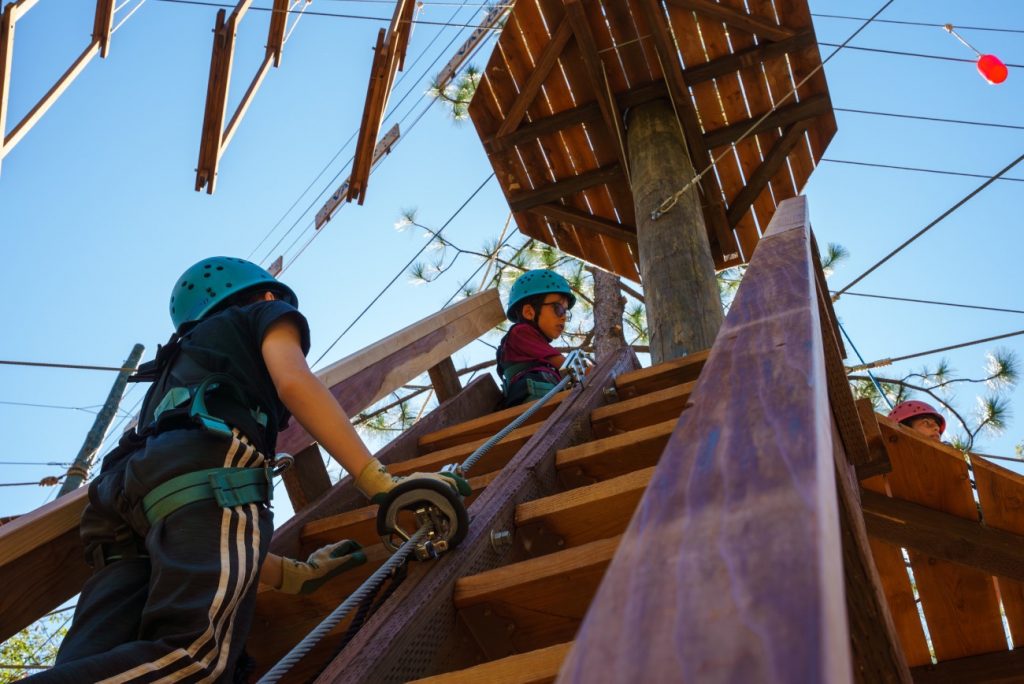 Two children wearing helmets and harnesses climb a wooden staircase toward a ropes course structure, preparing to participate in an outdoor adventure activity under a clear blue sky.