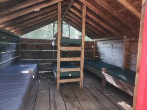 The image shows the inside of a rustic wooden cabin with three single beds and one bunk bed, all with thin mattresses. Sunlight filters through the wooden slats, creating a bright, airy atmosphere.