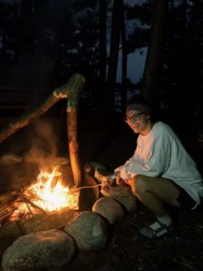 A person wearing glasses and a white sweatshirt squats by a campfire at night, roasting food on a stick. Large rocks surround the fire, and tall trees are visible in the dark background.