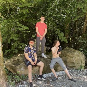 Three young men pose outdoors by large rocks and trees. One stands in a red shirt, the other two sit, one in sunglasses and floral shirt, the other shirtless, holding a black bag. Sunlight filters through the green foliage.