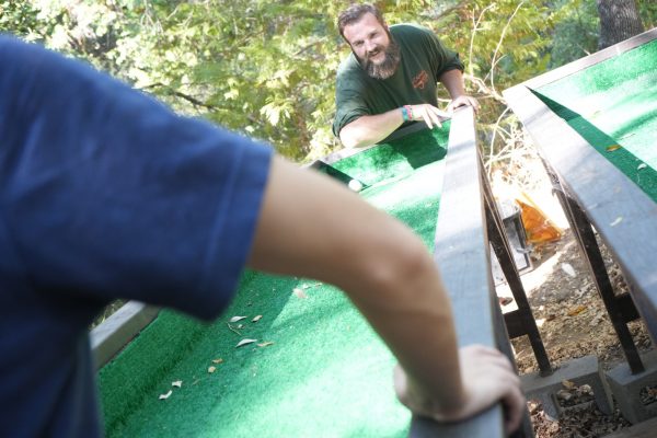 A man with a beard smiles while playing an outdoor game with a green ramp, as another person in a blue shirt stands in the foreground, both surrounded by trees.