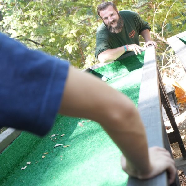 A man with a beard smiles while playing an outdoor game with a green ramp, as another person in a blue shirt stands in the foreground, both surrounded by trees.