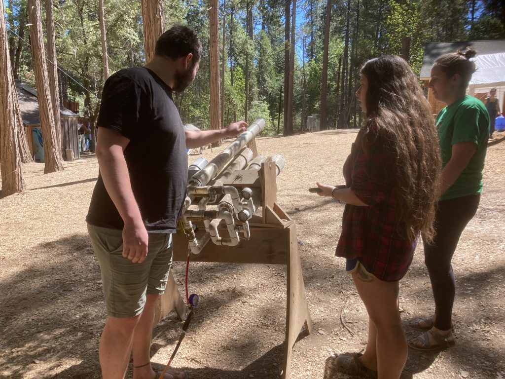 Three people stand outdoors in a wooded area examining a wooden and PVC pipe contraption on a stand, with sunlight streaming through tall trees around them.