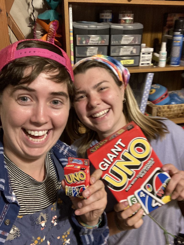 Two smiling people hold up decks of UNO cards, one regular size and one giant size, standing in a room with shelves of assorted items in the background.