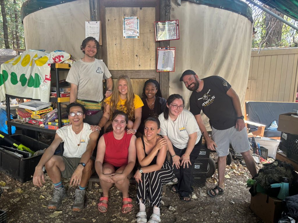 A group of eight people pose and smile together outdoors in front of a yurt. They are casually dressed and appear happy, surrounded by camping gear, games, and trees.